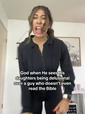 A young woman with light skin and brown hair is in a medium close-up, her face displaying a mix of amusement and disapproval. She wears a black zip-up jacket, and her mouth is open in a speaking position. The background is a simple living space with various decor elements.