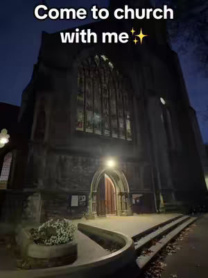 A photo of a large, gothic-style church at night. The church is made of stone and has a large arched window with stained glass. The entrance has an arched doorway, with a light illuminating the doorway. The sky is dark with some trees on the right side. In the foreground, there's a paved area. There is text overlaid toward the top of the image.