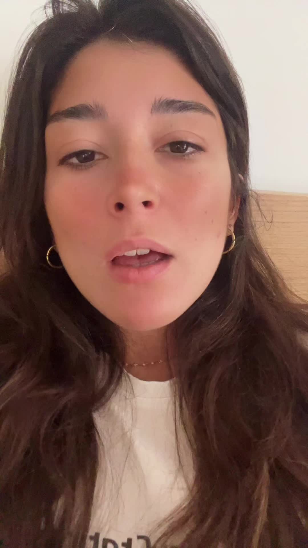 A close-up shot of a woman's face, angled slightly upward, with her eyes focused on the camera and her mouth slightly open. She has long, dark hair framing her face. Wavy hair flows down the sides. She's wearing a white shirt and gold earrings and a necklace. The background is a blurred neutral color. The lighting appears to be soft and natural.