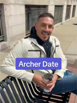 A man sits on a bench holding a coffee cup in front of a building with arched windows and a wrought-iron gate. The man is dressed in modern street style, with a white jacket, a black hooded garment underneath, and blue jeans. His face is centered, showing a smile, with a text overlay in a purple rectangular box, labeled 'Archer Date'.