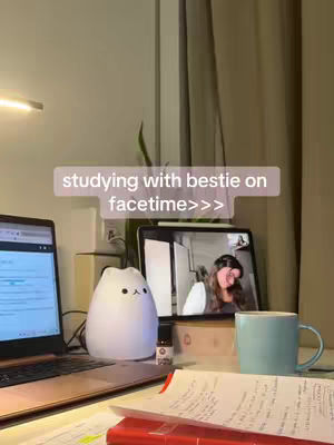 The image shows a well-lit desk with a laptop, a tablet displaying a video call, a lamp shaped like a rounded cat, a mug, and handwritten notes. The background is a neutral color, and the overall mood is one of focused productivity.