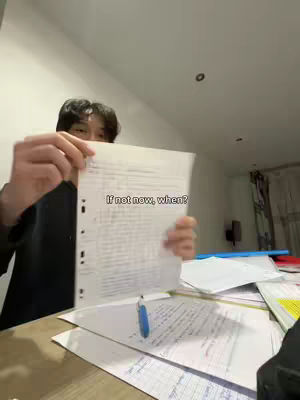 A person is holding a sheet of paper in front of them, with a handwritten note on it. The background reveals a cluttered desk with more papers and office supplies, suggesting a study or work environment. The person is partially visible, and the composition focuses on the message displayed on the held paper, suggesting motivation or a call to action.