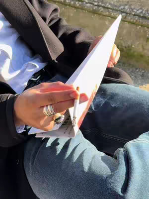 The cover image shows a person's lap with their hands holding a cigarette and a piece of paper, likely a menu or a piece of folded advertisement, while seated outside. The person wears a black coat on top of a white shirt and blue jeans. Several rings are visible on their fingers and gray smoke floats upward from the cigarette.