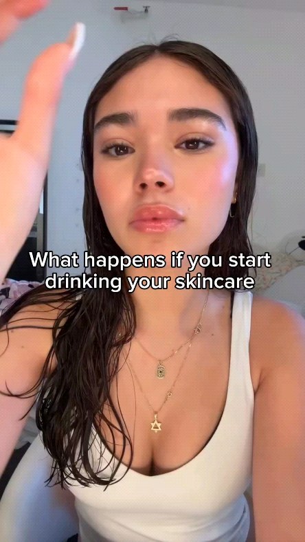 A selfie-style shot features a light-skinned woman with long dark hair, facing the camera. She's wearing a white tank top and several necklaces. A text overlay reads, "What happens if you start drinking your skincare". The background appears to be a neutral-toned room.