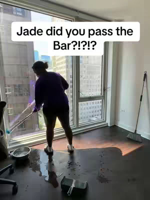 A person, viewed from the back, wearing a purple shirt and shorts, mops up a water spill in a room with large windows overlooking a city. The windows fill the background, and a sign that says "Jade did you pass the Bar?!?!?!", is placed above her head.