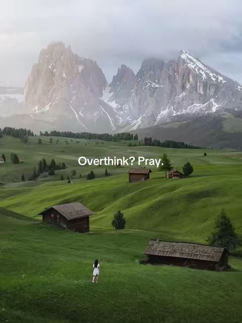 An image showcasing a scenic landscape featuring rolling green hills, scattered rustic wooden cabins, and a majestic mountain range in the background. A woman in a white dress is standing in the field. Text overlay on the image reads 'Overthink? Pray,' indicating a message-driven theme.