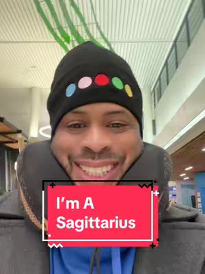 The cover image features a smiling Black man in a black beanie adorned with colorful dots. He has a travel pillow around his neck and is wearing a blue shirt. A red rectangular box with white text that reads, 'I'm A Sagittarius' is overlaid over the image. The background appears to be a public space with white ceiling panels.