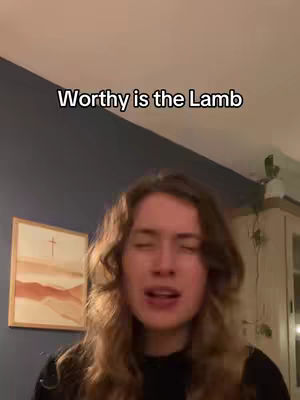 A person with long, wavy brown hair is singing, with their eyes closed, against a backdrop of a dark blue wall, a framed picture of a cross drawing, and a wooden cabinet. Text overlay says "Worthy is the Lamb".