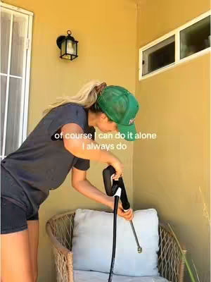 A woman in a green cap and casual clothes is outdoors, using a cleaning tool. The background is a beige wall with a window and a light fixture. A bench is in the frame.