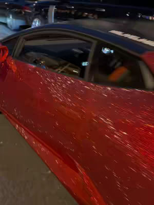 The cover image shows a portion of a shiny red supercar from the side, with focus on the car's exterior in the foreground. Other cars are blurred in the background. The composition is focused on the car's sleek design and vibrant color.