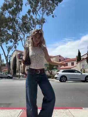 A woman stands on a city street, with a sunny sky behind her. She's dressed in a white lace tank top, blue jeans, and holds a bag and a drink. Buildings and cars are visible in the background.