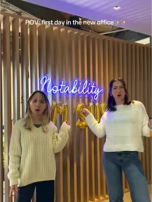 Two young women are posing in front of a wooden slatted wall. A neon sign reading "Notability" is behind them. They are holding gold number balloons that appear to spell out "2023".