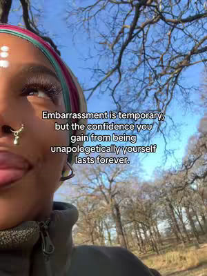 A close-up shot of a person's face, angled toward the viewer. The person has dark skin and is wearing a decorative head covering with colorful bands. Above their forehead, there are three white dots. A small golden nose ring hangs from their nostril. The background consists of trees and a blue sky.