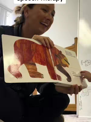 A woman is holding open a colorful children's book with a large bear illustration in one hand while smiling and looking off to the side. The book is facing towards the camera. A child's hand is visible, touching the book. The background includes part of a white wall with drawings on it and a blurred view of a room.