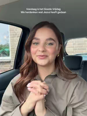 A woman sits in a car, facing the camera with a slight smile and her hands clasped together. She has long auburn hair, and is wearing a light-colored shirt. The interior of the car and a view of a brick building outside are visible.