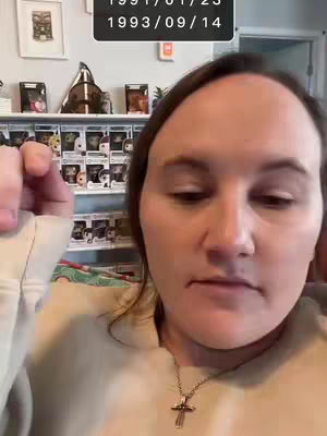 A woman is shown in a selfie-style shot. The background has a shelf with various pop-up figures and decorative objects. The woman is wearing a beige top and a cross necklace. Above her head, a black box overlays with two dates. The overall composition suggests a casual, at-home setting.