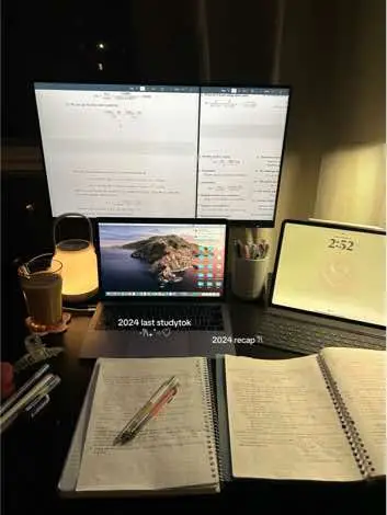 An aesthetically pleasing dark workspace setup at night, showing a laptop, multiple monitors with illuminated screens displaying what appears to be a study document, and open notebooks next to studying tools.
