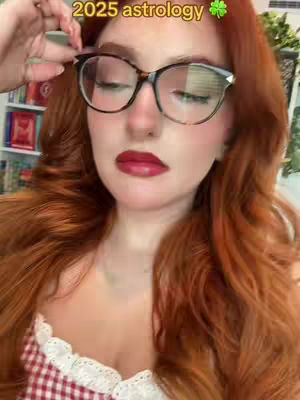 A woman with red hair and glasses is the central focus. The text '2025 astrology' is overlaid at the top, along with a green shamrock. The woman is wearing glasses and red lipstick. The background is blurred but includes what appears to be a bookshelf.