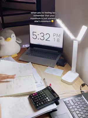 The image shows a workspace with a laptop displaying 5:32 PM, a calculator, and study materials. A soft toy penguin sits to the left, and a bright desk lamp illuminates the scene. The text overlayed says motivation message about lazy feeling and minimum effort.