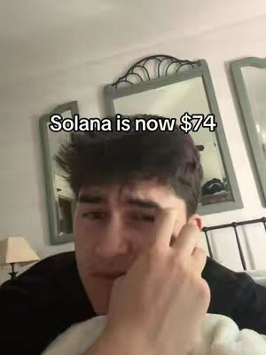 A selfie-style video shows a young man in a room with mirrors. The overlay text states, 'Solana is now $74.' The man is looking slightly off-camera with a hand to his face, with a look of mild concern.