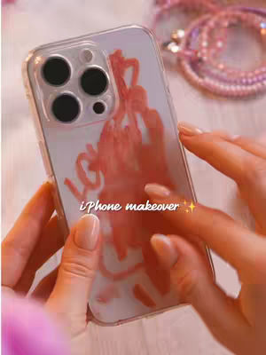A close-up shot showcasing a person holding a phone with a customized pink background displayed. The phone is enclosed in a clear case, and the person's hands and portions of a pink beaded accessory are also visible. The background is softly illuminated.