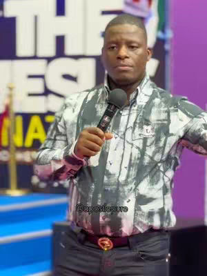 A man in a patterned shirt holds a microphone, standing in front of a blurred background with text. The lighting is bright, and the man has a serious expression.