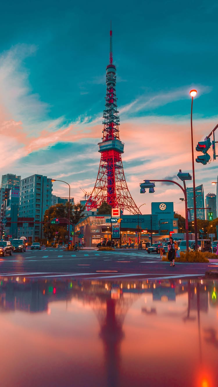 The image shows the Tokyo Tower in Japan, seen from a street view on a cloudy day at sunset. The tower is the central element, with its red and white lattice structure standing tall against an orange and blue sky. Buildings and city lights are visible along the edges of the image, and the street reflects the lights below, creating a serene and vibrant atmosphere.