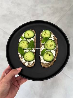 An overhead shot showcases a black plate centered on a light gray surface. The plate holds two open-faced sandwiches, composed of sliced cucumber, white spread, and green sprigs, likely mint, atop toasted bread. A hand, adorned with a ring, gently cradles the plate, suggesting the meal is prepared and held for the shot. The backdrop appears to be a plain, off-white surface, contributing to the clean aesthetic focus.