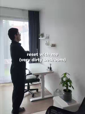 A person stands in a room with a standing desk, a window, and a plant, looking toward the window. Text overlayed indicates a bedroom reset.