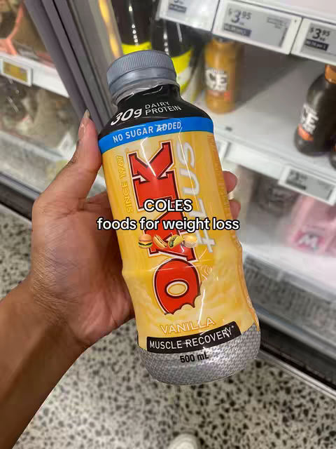 A hand holding a bottle of OK vanilla muscle recovery drink inside of a grocery store setting. The bottle is brightly colored and has a black cap; the background shows shelves and other products.