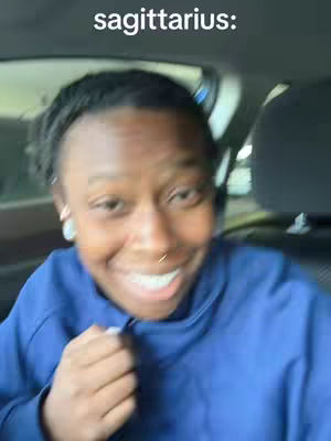A selfie-style shot taken from inside of a car shows a woman with short, dark hair smiling widely at the camera. The text overlay at the top reads 'sagittarius:'. The woman is wearing a blue shirt and a nose ring.