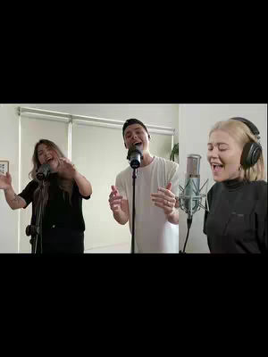 Three people are singing into microphones in a studio setting. The person in the middle is a male, and the other two are female singers. They a dressed in contrasting black and white shirts and are in a studio.