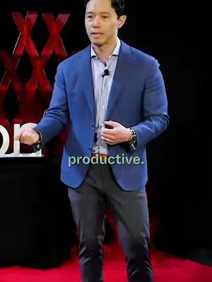 A man in a blue blazer and gray pants stands on a stage, presenting. The background appears to be a red and black stage design. Text is overlaid at the bottom of the image.