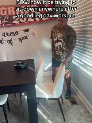 A man in a camo t-shirt and black shorts struggles to sit down on a chair after a leg day workout. The image is captured in an indoor setting with natural light and includes a table with an ashtray, blinds, and a decorative banner. 