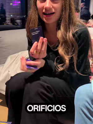 A woman is holding a deck of cards while talking. Below her, there is a white text bubble with the text 'ORIFICIOS'. The background seems to be a room with a window looking out onto city lights.