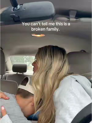 A woman with long blonde hair is sitting in the front seat of a car, embracing a person whose face is partially visible, resting it on her shoulder. The interior of the car, including the rearview mirror, is visible, with text overlaid on the image.
