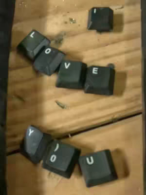 The image features a collection of black keyboard keys arranged to spell out "I LOVE YOU". The keys are scattered across a wooden surface, possibly a table or desk, with various pieces of keyboard key caps. The layout gives a sense of a casual and personal message. The arrangement makes the text readable, with the keys slightly tilted in different directions. The wooden surface has a natural wood grain. The lighting appears diffused. The key caps stand out against the wooden background.