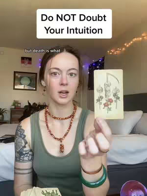 A woman with fair skin gazes directly at the camera, holding a tarot card in her right hand. Above her head is a white rectangle with black text. The background features a bedroom setting, with fairy lights and a bed.