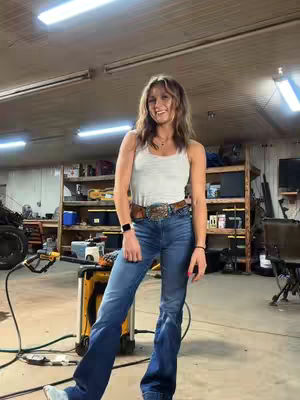 A woman stands in a workshop setting, wearing a white tank top, jeans, and a brown belt with a detailed buckle. She smiles at the camera. The background includes shelving with various items and tools, and bright overhead lighting.