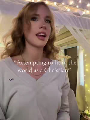 A young woman with light brown, wavy hair is centered in the frame, looking towards the viewer with a slightly open mouth as if mid-sentence. She is wearing a light-colored sweater, and her face is lightly made up. Behind her is a soft, blurry background, which includes fairy lights and a sheer white curtain, suggesting an indoor setting with a festive mood. The image is well-lit, with the focus on the woman's face.
