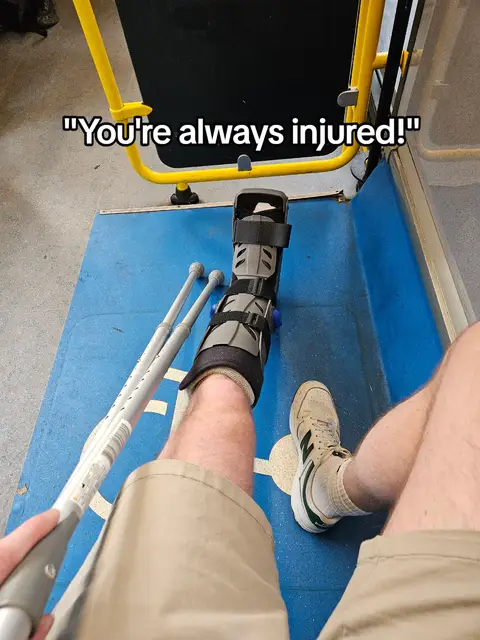 A first-person perspective shows a person in a wheelchair space, wearing a leg brace. The person's leg with a brace and crutches is visible. Above, text states, "You're always injured!"