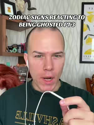 A selfie-style video featuring a man with fair skin, brown eyes, and short, dark hair. He is wearing a dark green t-shirt with text and holding a white earphone cord. Behind him are a wooden shelf with various items and a framed picture.