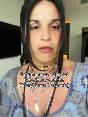 A woman with dark hair and fair skin looks directly at the camera. She wears multiple necklaces. The background includes a glimpse of a room with a television and a closed door.