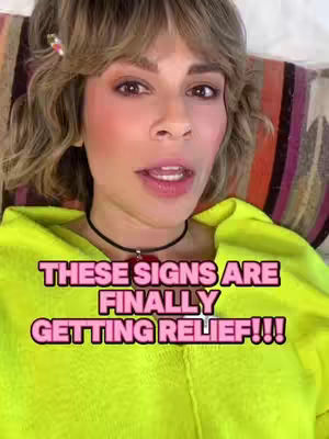 A woman with short, wavy blonde hair is centered in the frame, looking directly at the camera with a slight smile and open mouth. She wears a bright yellow hoodie and a delicate black choker. Text overlaid in a pink and black box reads "THESE SIGNS ARE FINALLY GETTING RELIEF!!!". The background features a colorful, striped pillow.