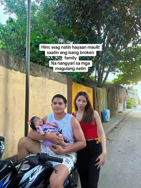 A family of three poses on a street with a motorcycle. A man in a white tank top and shorts holds a baby, while a woman in a red top and black pants stands nearby. Text with a white background is overlaid on the top-left of the image.