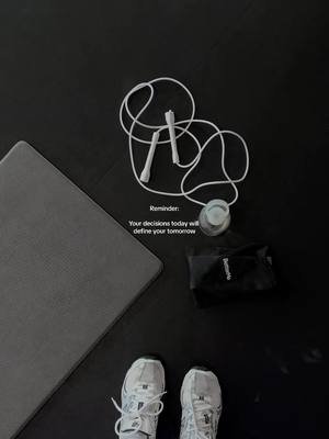 The image is a top-down view of a workout setting. A grey yoga mat is in the upper left, angled. In the center, a white jump rope is coiled. A small black bag and a water bottle are next to the jump rope. At the bottom of the image are the tops of a pair of white sneakers. The floor is black.