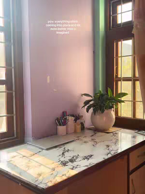 The image showcases a well-lit study space with a window on the right and a window on the left. A desk with a marble-patterned surface fills the foreground. There are various office supplies and a plant in a white pot on the desk. The wall is painted in a pastel color, and text which is added with the overlay on the wall displays in white font. The scene is calm and inviting.