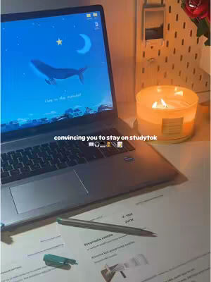A well-lit interior shot featuring a laptop, a lit candle, and study materials. The laptop displays a wallpaper of a whale swimming in a blue sky with a moon and stars. A pen rests on a sheet of paper, and a stapler and other stationery items are visible next to the laptop. The candle in a glass jar is positioned to the right, casting a warm glow.