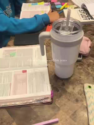 A person wearing a blue sweatshirt is sitting at a table with a Bible and other books open on it. A large white tumbler with a handle and straw is to the right of the open book. The table is a mottled pattern in light brown and tan.