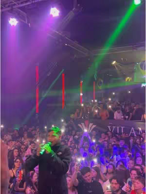 A man is on stage, holding a microphone and singing in front of a large crowd in a club setting, surrounded by vibrant stage lighting, spotlights, and a sea of people holding up their phones. The layout and focus are on the performer and the energy of the event.